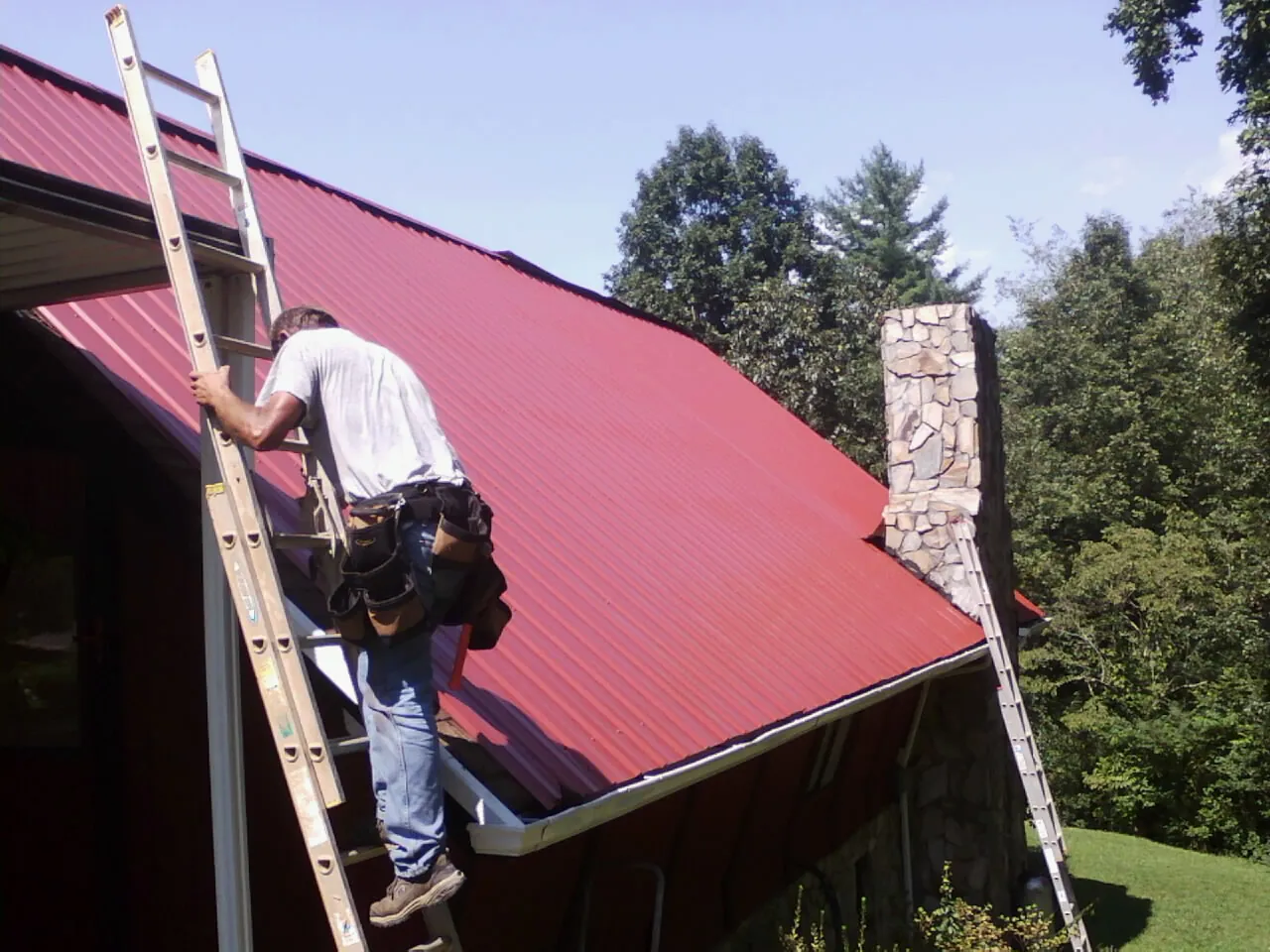 Roofer on a ladder beside a red metal roof and stone chimney — Soffit & Fascia Repair by Roofing Brothers in Tilton